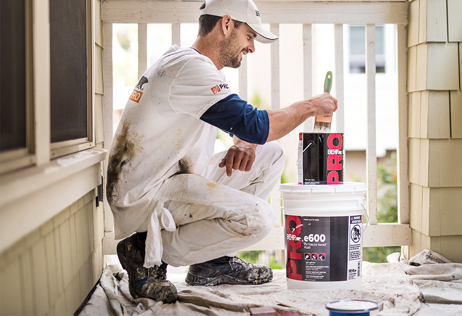 Pro Contractor wearing a cap and shirt with Behr logo dipping a paint brush on a 1 gallon of BEHR PRO e600 paint can on a porch that he is going to paint.